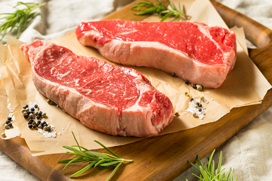 Two raw NY strip steaks on butcher paper with salt, pepper, and rosemary sprigs. 