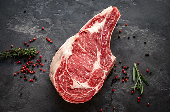 A raw tomahawk steak, also known as a bone-in ribeye on a gray background with red peppercorns scattered.