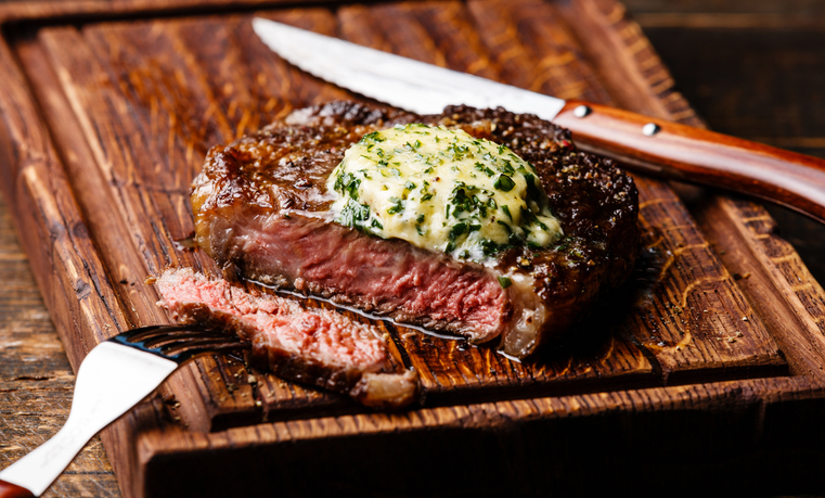 A grilled ribeye with seasoned butter and utensils on a wooden cutting board.